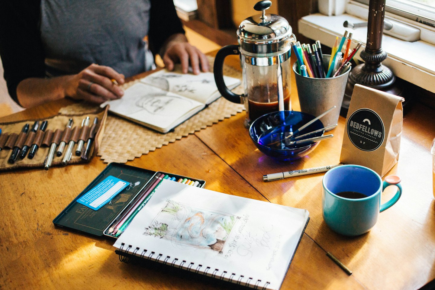 Hands sketching in a notebook on a wooden table with art supplies.