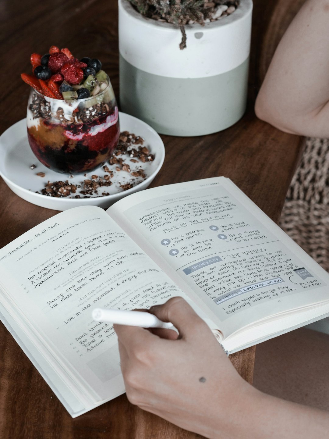 Hand writing in journal next to fruit parfait on wooden table.
